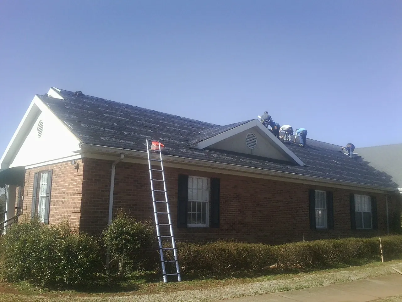 Crew tearing shingles off a brick ranch roof with a ladder set — Emergency Roof Repair work by our crew in Petoskey
