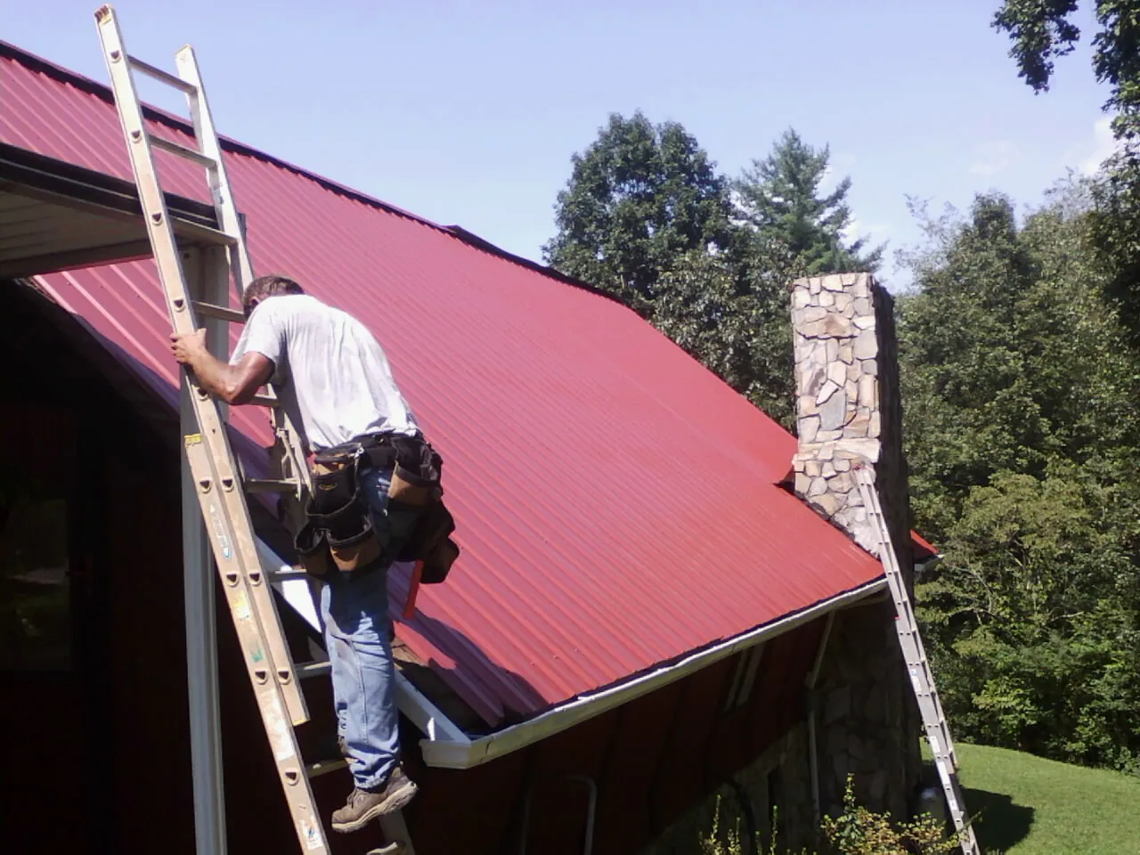 Roofer on a ladder beside a red metal roof and stone chimney — Soffit & Fascia Repair by Roofing Brothers in Petoskey